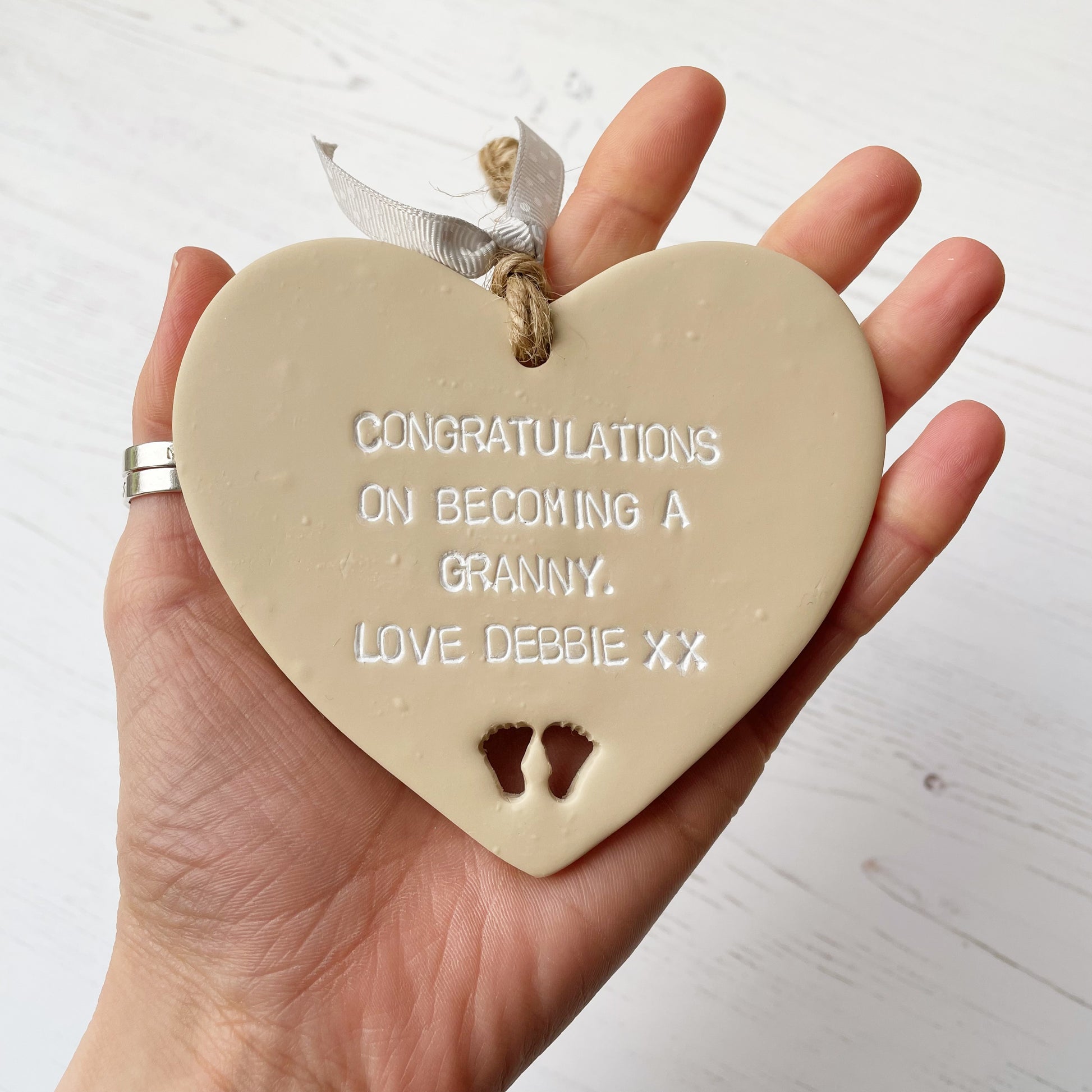 Beige clay hanging heart with baby feet cut out of the bottom and white personalisation, the heart is personalised with CONGRATULATIONS ON BECOMING A GRANNY. LOVE DEBBIE XX