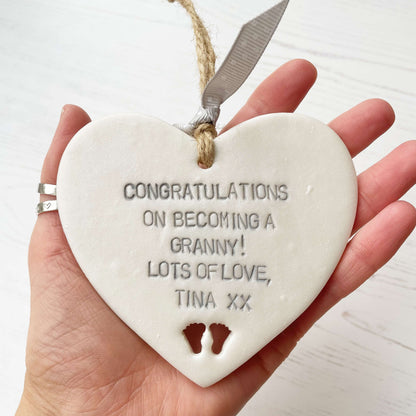 Pearlised white clay hanging heart with baby feet cut out of the bottom and grey personalisation, the heart is personalised with CONGRATULATIONS ON BECOMING A GRANNY! LOTS OF LOVE TINA XX