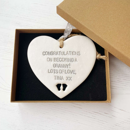Pearlised white clay hanging heart with baby feet cut out of the bottom and grey personalisation, the heart is personalised with CONGRATULATIONS ON BECOMING A GRANNY! LOTS OF LOVE TINA XX