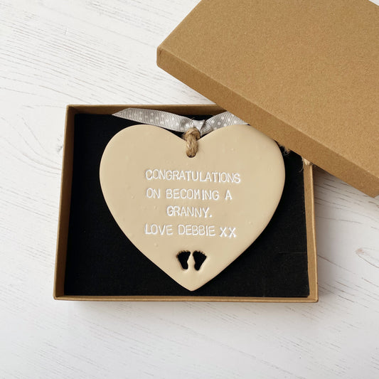 Beige clay hanging heart with baby feet cut out of the bottom and white personalisation, the heart is personalised with CONGRATULATIONS ON BECOMING A GRANNY. LOVE DEBBIE XX