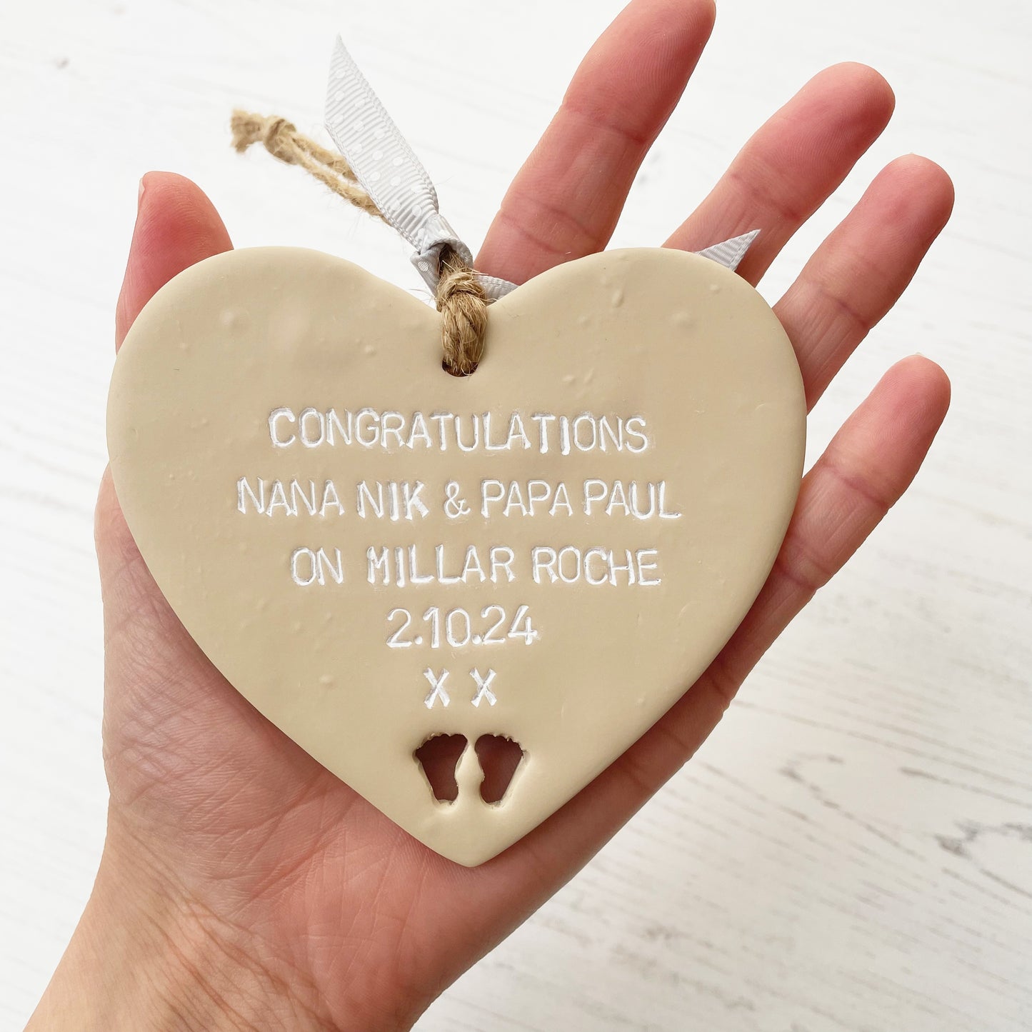 Beige clay hanging heart with baby feet cut out of the bottom and white personalisation, the heart is personalised with CONGRATULATIONS NANA NIK & PAPA PAUL ON MILLAR ROCHE 2.10.24 XX