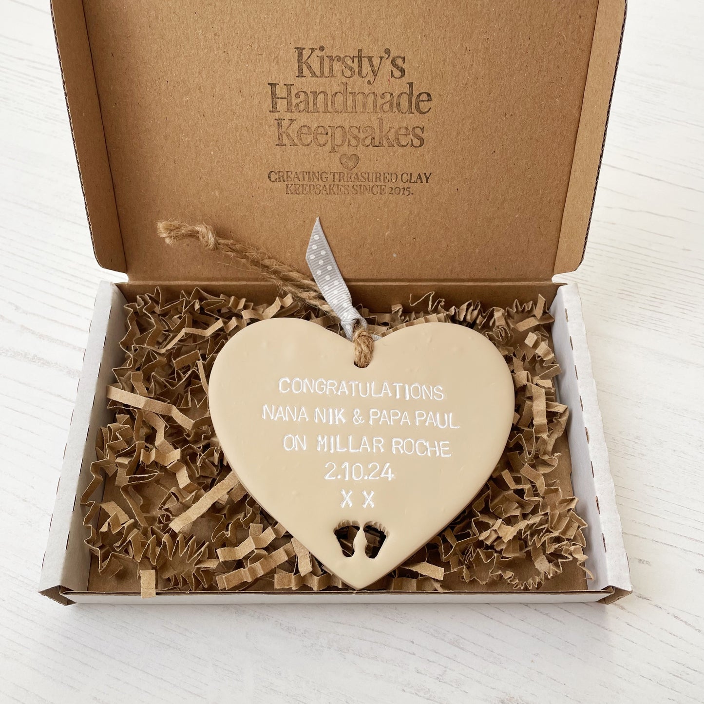 Beige clay hanging heart with baby feet cut out of the bottom and white personalisation, the heart is personalised with CONGRATULATIONS NANA NIK & PAPA PAUL ON MILLAR ROCHE 2.10.24 XX