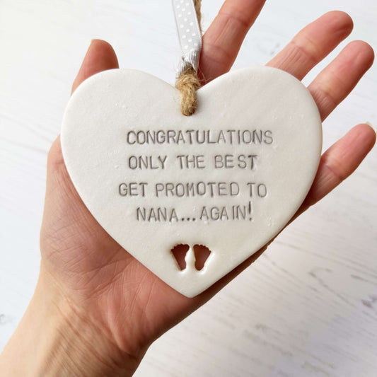 Pearlised white clay hanging heart with baby feet cut out of the bottom and grey personalisation, the heart is personalised with CONGRATULATIONS ONLY THE BEST GET PROMOTED TO NANA… AGAIN!