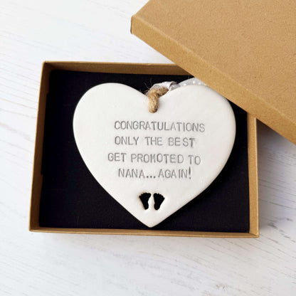 Pearlised white clay hanging heart with baby feet cut out of the bottom and grey personalisation, the heart is personalised with CONGRATULATIONS ONLY THE BEST GET PROMOTED TO NANA… AGAIN!