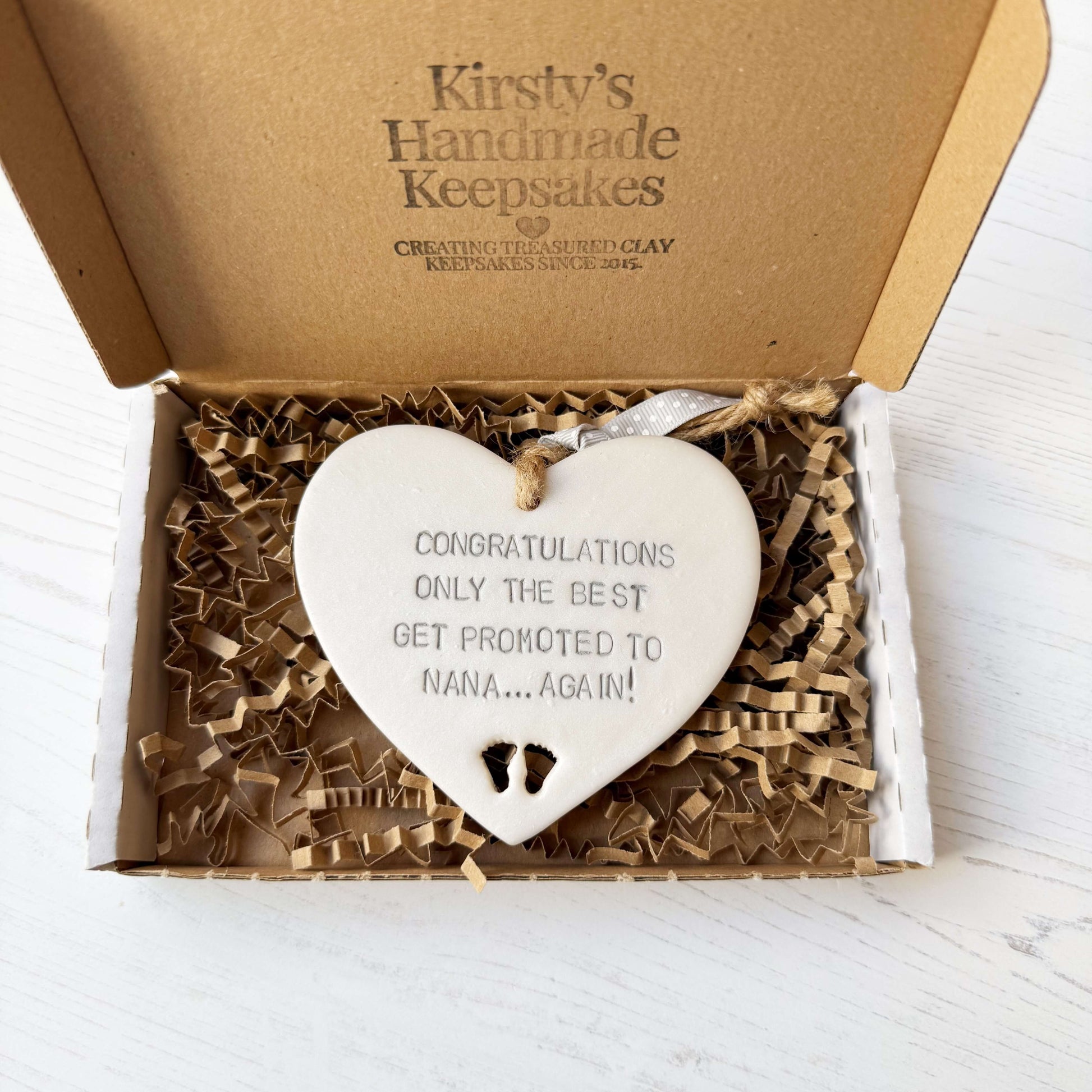 Pearlised white clay hanging heart with baby feet cut out of the bottom and grey personalisation, the heart is personalised with CONGRATULATIONS ONLY THE BEST GET PROMOTED TO NANA… AGAIN!