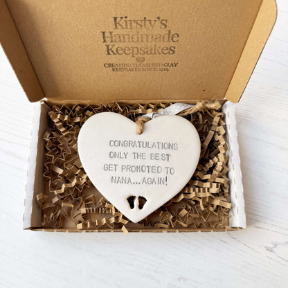 Pearlised white clay hanging heart with baby feet cut out of the bottom and grey personalisation, the heart is personalised with CONGRATULATIONS ONLY THE BEST GET PROMOTED TO NANA… AGAIN!
