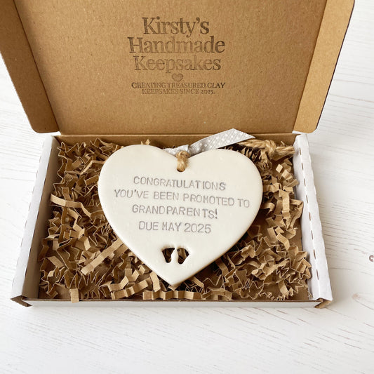 Pearlised white clay hanging heart with baby feet cut out of the bottom and grey personalisation, the heart is personalised with CONGRATULATIONS YOU’VE BEEN PROMOTED TO GRANDPARENTS! DUE MAY 2025