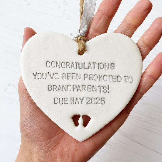 Pearlised white clay hanging heart with baby feet cut out of the bottom and grey personalisation, the heart is personalised with CONGRATULATIONS YOU’VE BEEN PROMOTED TO GRANDPARENTS! DUE MAY 2025