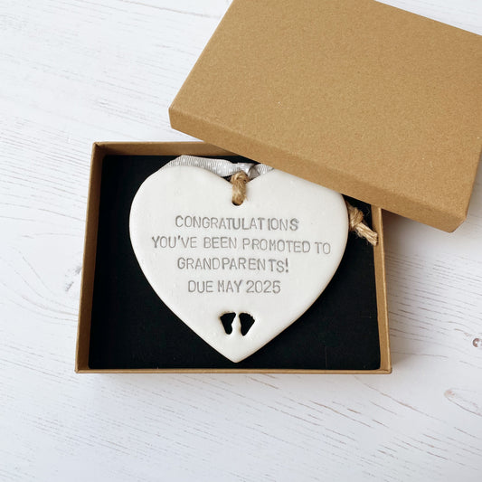 Pearlised white clay hanging heart with baby feet cut out of the bottom and grey personalisation, the heart is personalised with CONGRATULATIONS YOU’VE BEEN PROMOTED TO GRANDPARENTS! DUE MAY 2025