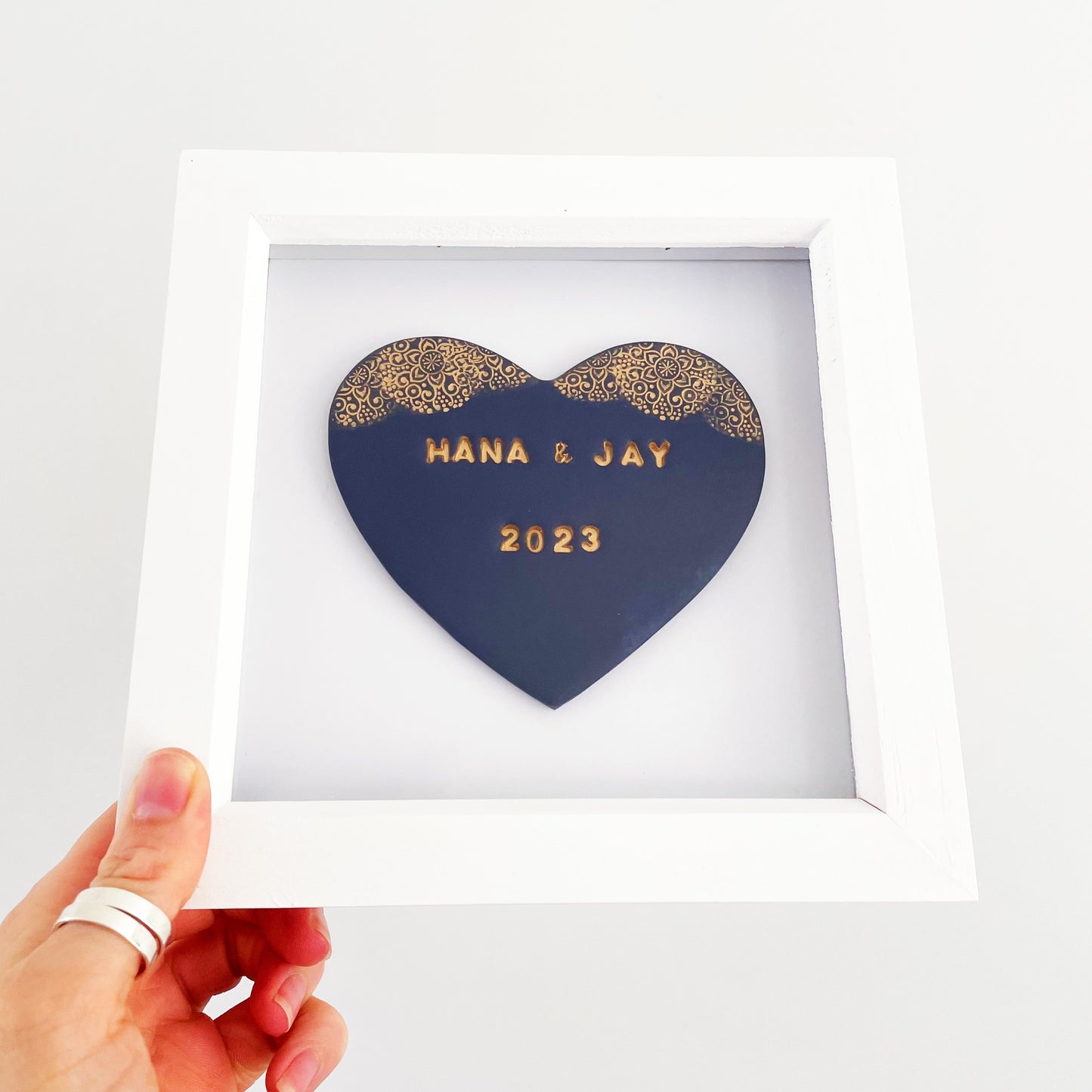Personalised framed heart gift, dark blue clay heart with a gold lace edge at the top of the heart in a white box frame, the heart is personalised with HANA & JAY 2023