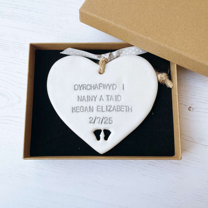 Pearlised white clay hanging heart with baby feet cut out of the bottom and grey personalisation, the heart is personalised with DYRCHAFWYD I NAINY & TAID MEGAN ELIZABETH 2/7/25