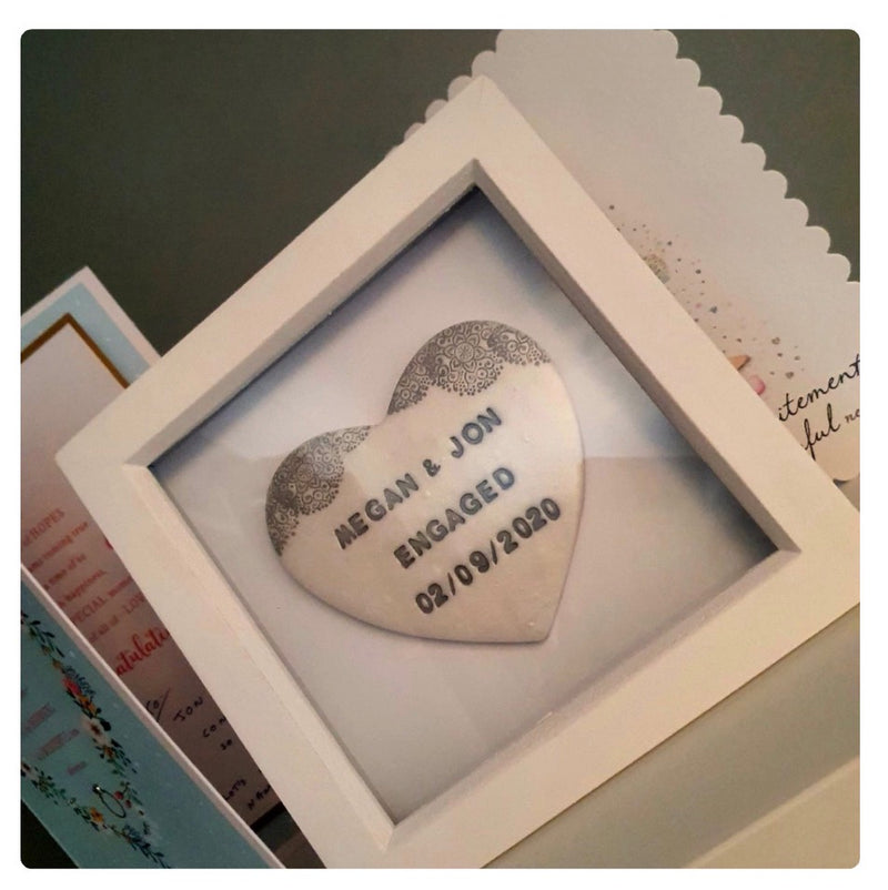 A white wooden shadow box frame displaying a pearlised white polymer clay heart with a grey lace edge and personalised text reading 'MEGAN & JON ENGAGED 02/09/2020'.