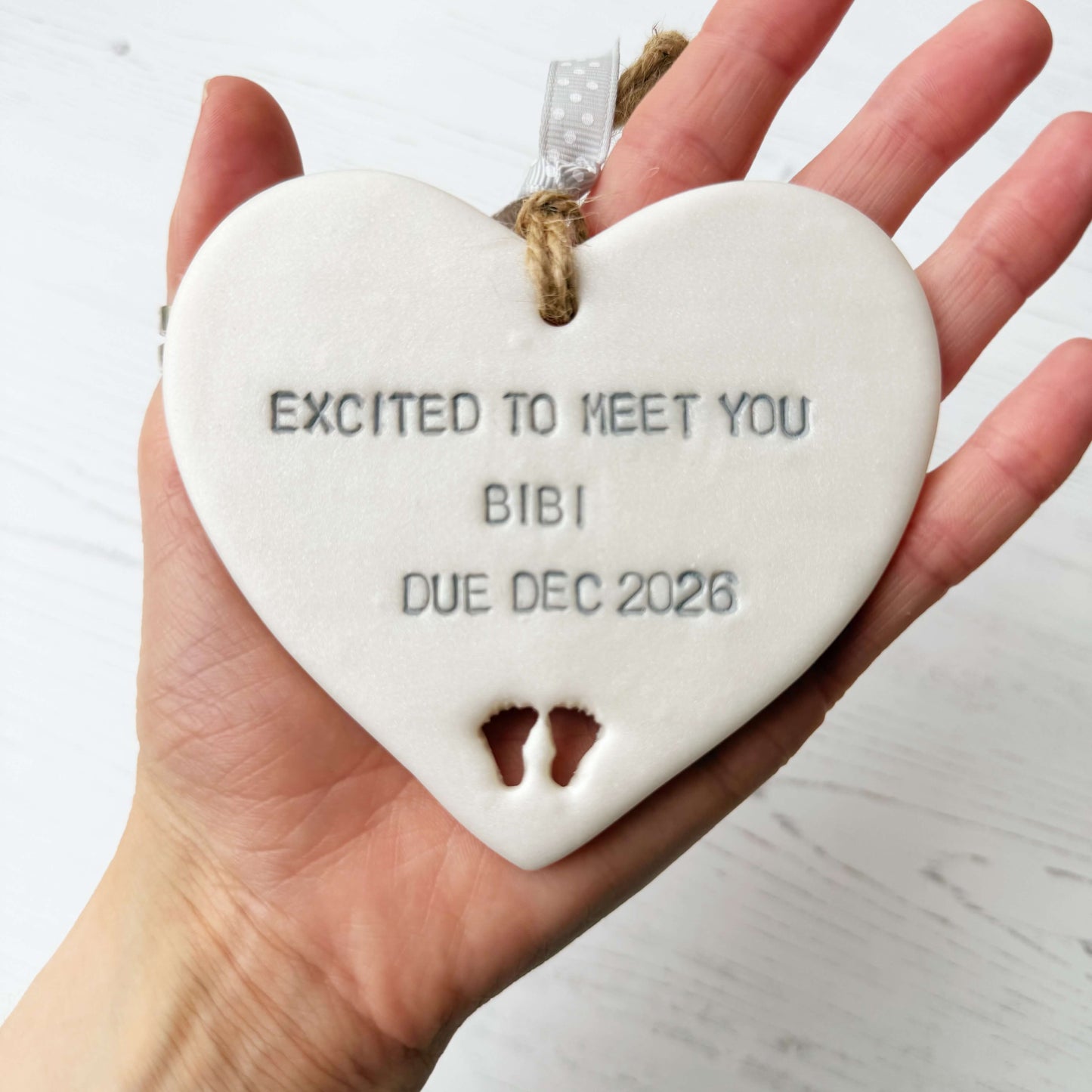 Pearlised white clay hanging heart with baby feet cut out of the bottom and grey personalisation, the heart is personalised with EXCITED TO MEET YOU BIBI DUE DEC 2026