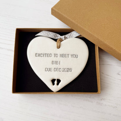 Pearlised white clay hanging heart with baby feet cut out of the bottom and grey personalisation, the heart is personalised with EXCITED TO MEET YOU BIBI DUE DEC 2026