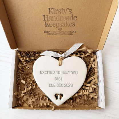 Pearlised white clay hanging heart with baby feet cut out of the bottom and grey personalisation, the heart is personalised with EXCITED TO MEET YOU BIBI DUE DEC 2026