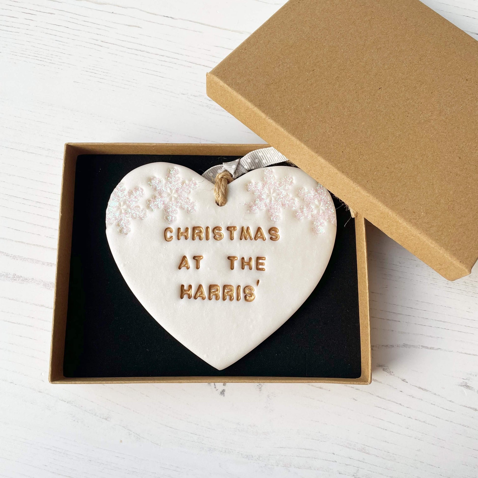 Personalised family Christmas heart ornament, pearlised white clay with CHRISTMAS AT THE HARRIS’ painted gold, decorated with 2 iridescent glitter snowflakes on either side of the top of the heart