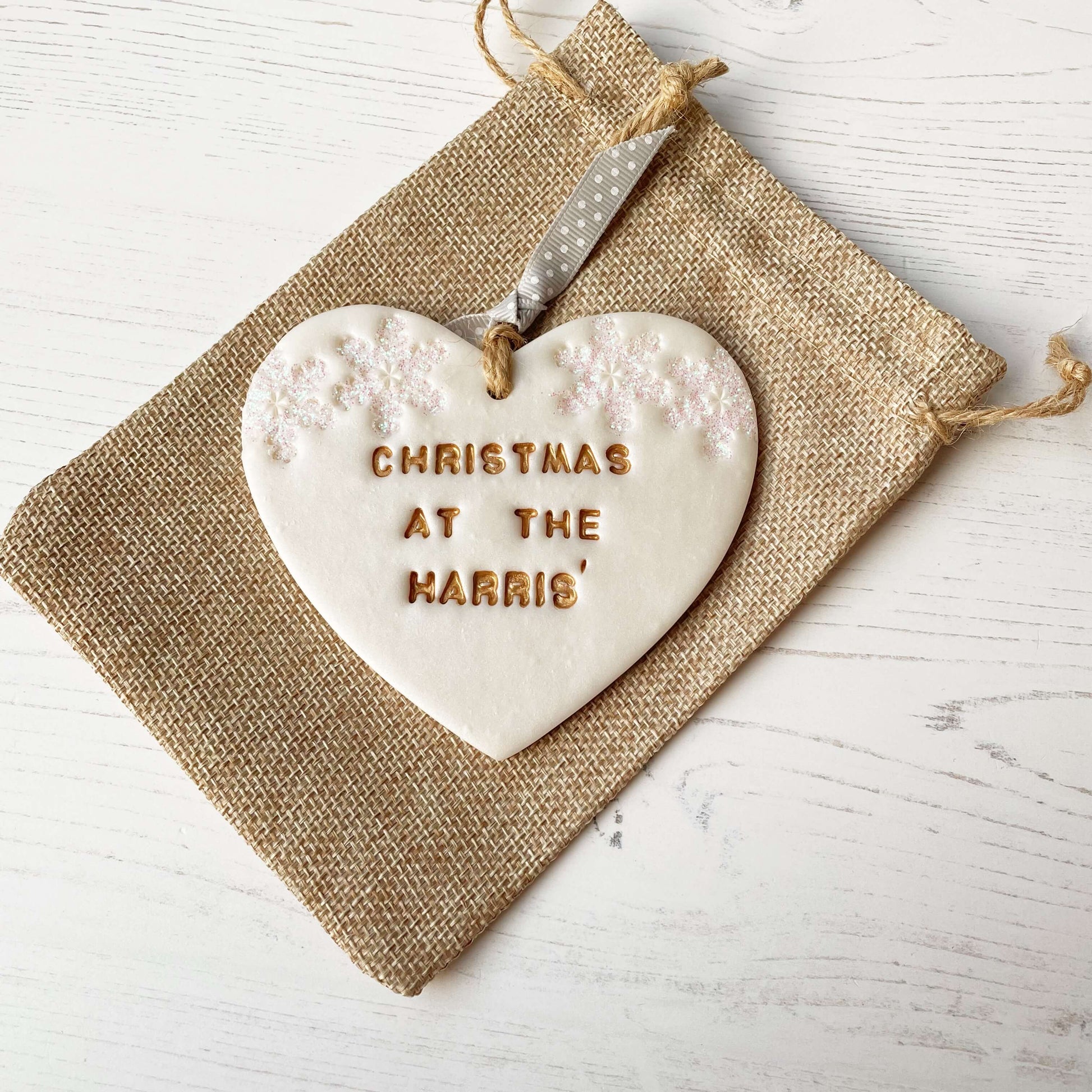 Personalised family Christmas heart ornament, pearlised white clay with CHRISTMAS AT THE HARRIS’ painted gold, decorated with 2 iridescent glitter snowflakes on either side of the top of the heart