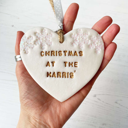 Personalised family Christmas heart ornament, pearlised white clay with CHRISTMAS AT THE HARRIS’ painted gold, decorated with 2 iridescent glitter snowflakes on either side of the top of the heart