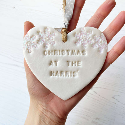 Personalised family Christmas heart ornament, pearlised white clay with CHRISTMAS AT THE HARRIS’ (text not painted), decorated with 2 iridescent glitter snowflakes on either side of the top of the heart