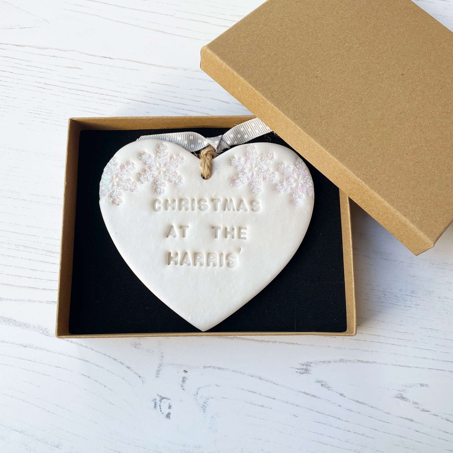 Personalised family Christmas heart ornament, pearlised white clay with CHRISTMAS AT THE HARRIS’ (text not painted), decorated with 2 iridescent glitter snowflakes on either side of the top of the heart
