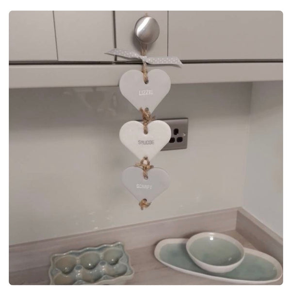 Three white and grey hearts hanging from a silver door handle with twine, labeled 'LOZIE', 'SPLURGE', and 'SCAMPI' in small black text. The background shows a kitchen wall and countertop with a ceramic egg tray and dish.