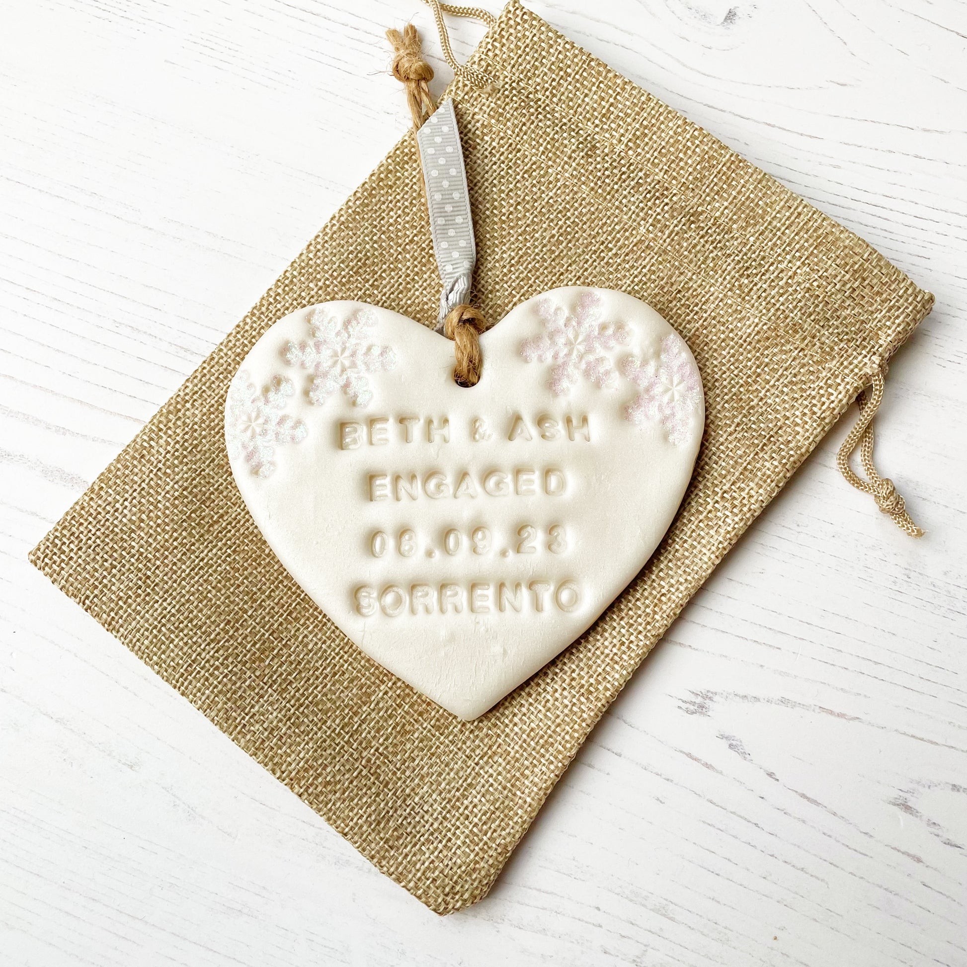 Personalised first Christmas engaged heart ornament, pearlised white clay with BETH & ASH ENGAGED 08.09.23 SORRENTO (text not painted), decorated with 2 iridescent glitter snowflakes on either side of the top of the heart