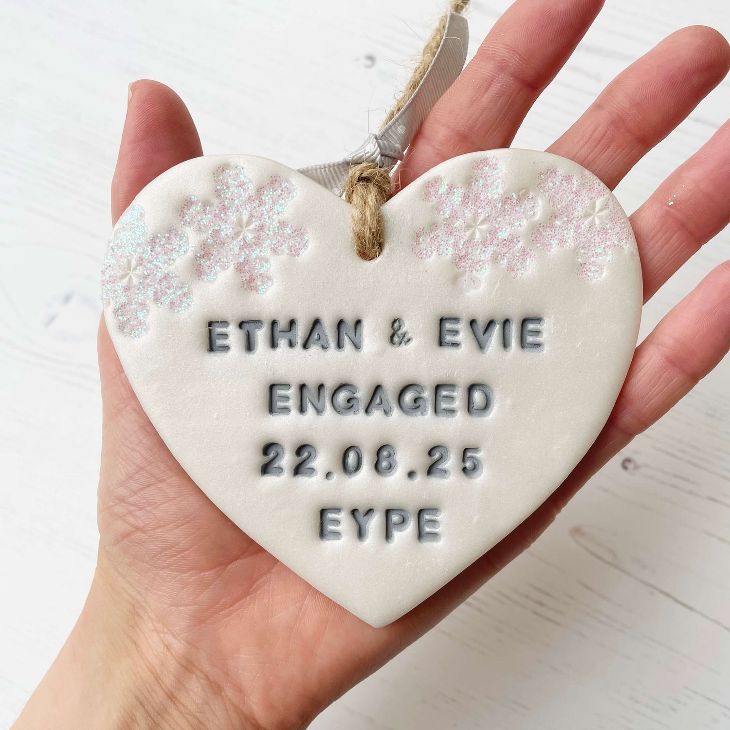 Personalised first Christmas engaged heart ornament, pearlised white clay with ETHAN & EVIE ENGAGED 22.08.25 EYPE painted grey, decorated with 2 iridescent glitter snowflakes on either side of the top of the heart