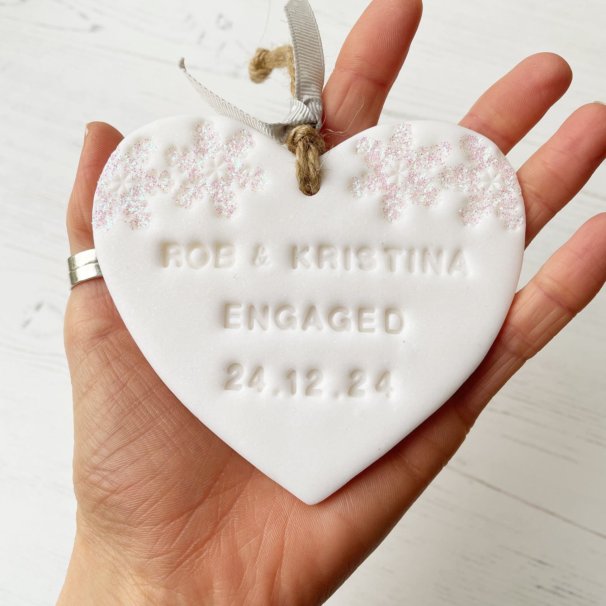 Personalised first Christmas engaged heart ornament, glitter white clay with ROB & KRISTINA ENGAGED 24.12.24 (text not painted), decorated with 2 iridescent glitter snowflakes on either side of the top of the heart