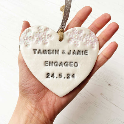 Personalised first Christmas engaged heart ornament, pearlised white clay with TAMSIN & JAMIE ENGAGED 24.5.24 painted grey, decorated with 2 iridescent glitter snowflakes on either side of the top of the heart