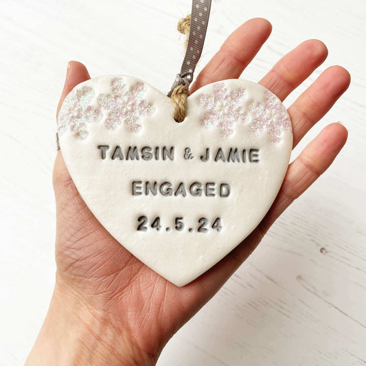 Personalised first Christmas engaged heart ornament, pearlised white clay with TAMSIN & JAMIE ENGAGED 24.5.24 painted grey, decorated with 2 iridescent glitter snowflakes on either side of the top of the heart