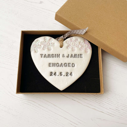 Personalised first Christmas engaged heart ornament, pearlised white clay with TAMSIN & JAMIE ENGAGED 24.5.24 painted grey, decorated with 2 iridescent glitter snowflakes on either side of the top of the heart