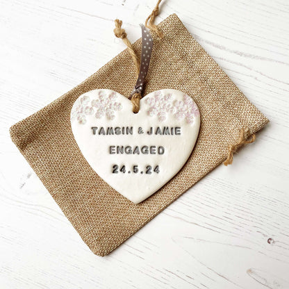 Personalised first Christmas engaged heart ornament, pearlised white clay with TAMSIN & JAMIE ENGAGED 24.5.24 painted grey, decorated with 2 iridescent glitter snowflakes on either side of the top of the heart