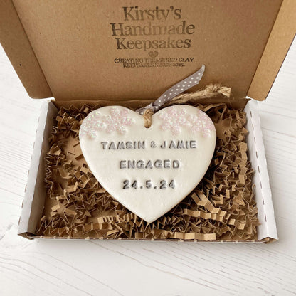 Personalised first Christmas engaged heart ornament, pearlised white clay with TAMSIN & JAMIE ENGAGED 24.5.24 painted grey, decorated with 2 iridescent glitter snowflakes on either side of the top of the heart