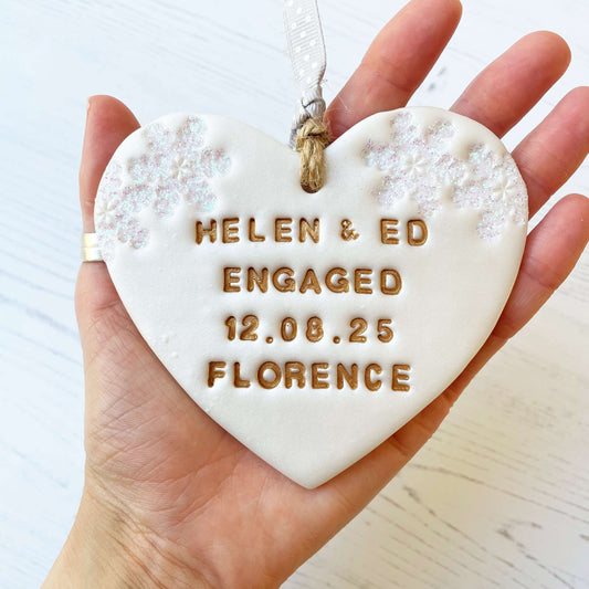 Personalised first Christmas engaged heart ornament, pearlised white clay with HELEN & ED ENGAGED 12.08.25 FLORENCE painted gold, decorated with 2 iridescent glitter snowflakes on either side of the top of the heart