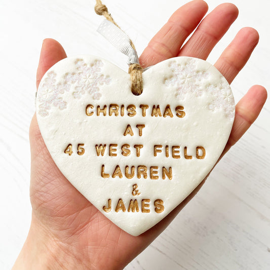 Personalised first Christmas in our new home heart ornament, pearlised white clay with CHRISTMAS AT 45 WEST FIELD LAUREN & JAMES painted gold, decorated with 2 iridescent glitter snowflakes on either side of the top of the heart