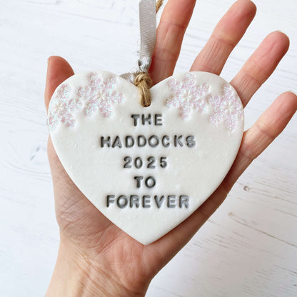 Personalised first Christmas married heart ornament, pearlised white clay personalised in grey with THE HADDOCKS 2025 TO FOREVER decorated with 2 iridescent glitter snowflakes on either side of the top of the heart