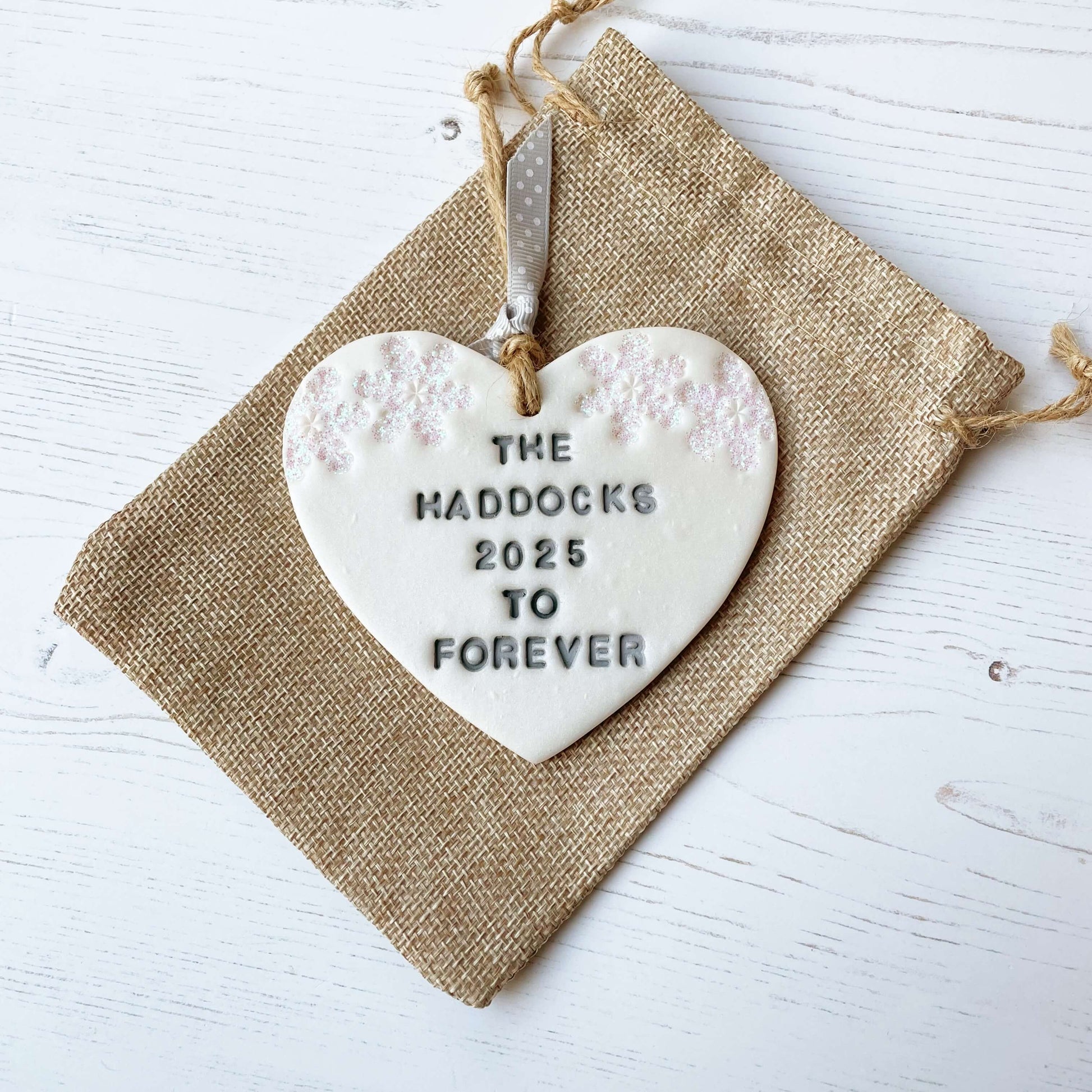 Personalised first Christmas married heart ornament, pearlised white clay personalised in grey with THE HADDOCKS 2025 TO FOREVER decorated with 2 iridescent glitter snowflakes on either side of the top of the heart