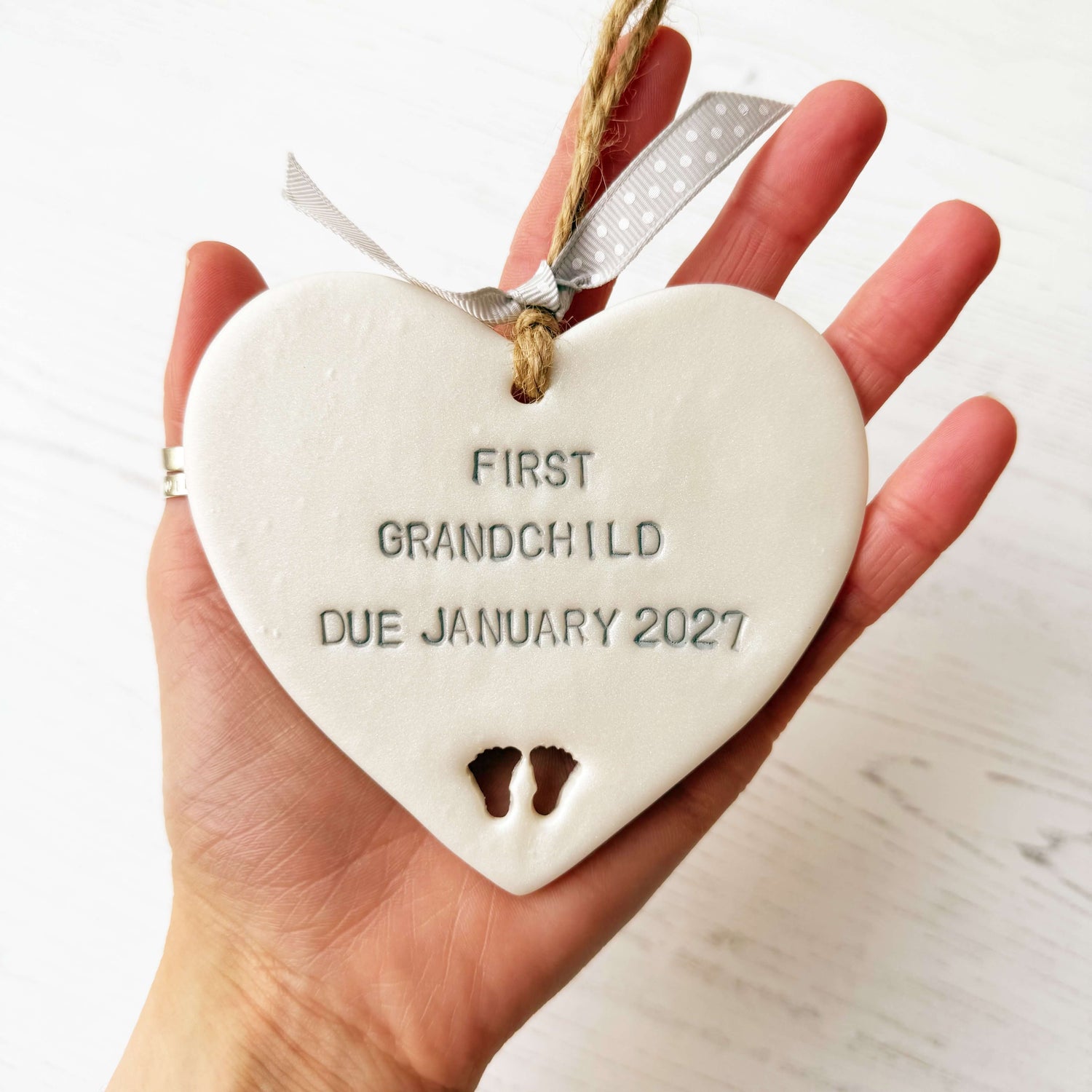 Pearlised white clay hanging heart with baby feet cut out of the bottom and grey personalisation, the heart is personalised with FIRST GRANDCHILD DUE JANUARY 2027