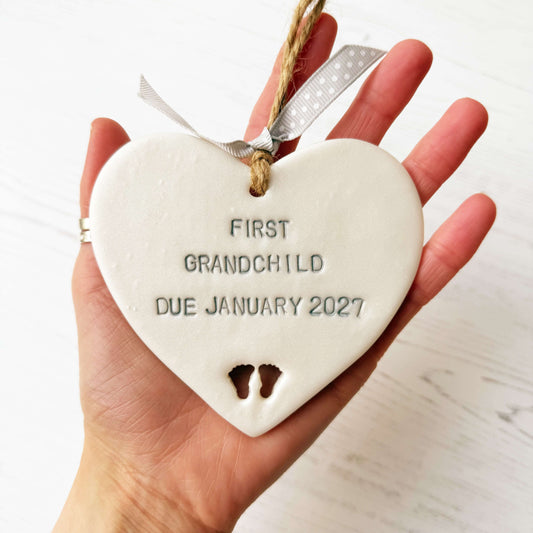 Pearlised white clay hanging heart with baby feet cut out of the bottom and grey personalisation, the heart is personalised with FIRST GRANDCHILD DUE JANUARY 2027
