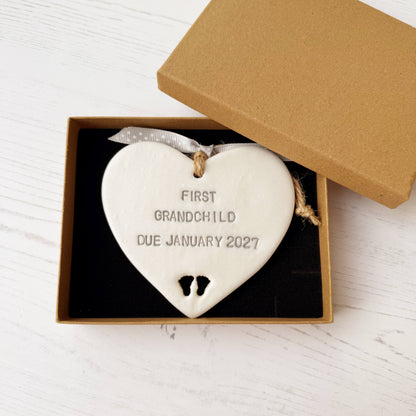 Pearlised white clay hanging heart with baby feet cut out of the bottom and grey personalisation, the heart is personalised with FIRST GRANDCHILD DUE JANUARY 2027