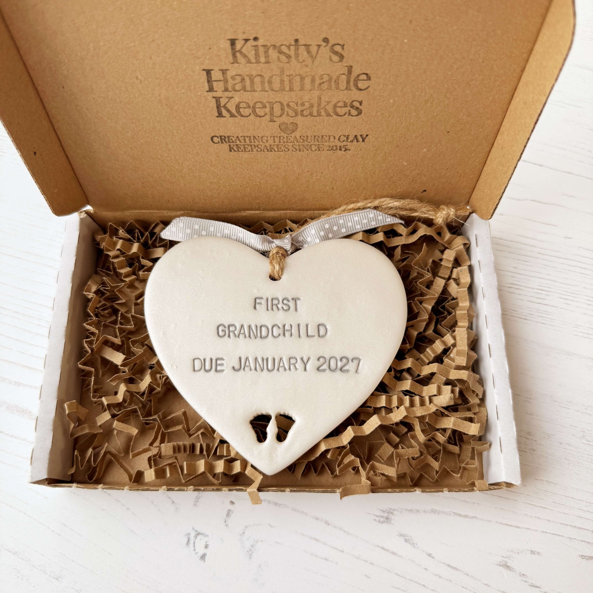 Pearlised white clay hanging heart with baby feet cut out of the bottom and grey personalisation, the heart is personalised with FIRST GRANDCHILD DUE JANUARY 2027