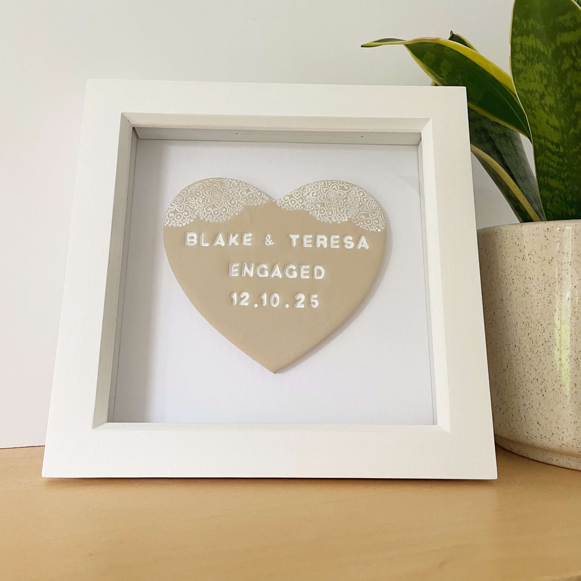 Personalised framed engagement gift, beige clay heart with a white lace edge at the top of the heart in a white box frame, the heart is personalised with BLAKE & TERESA ENGAGED 12.10.25