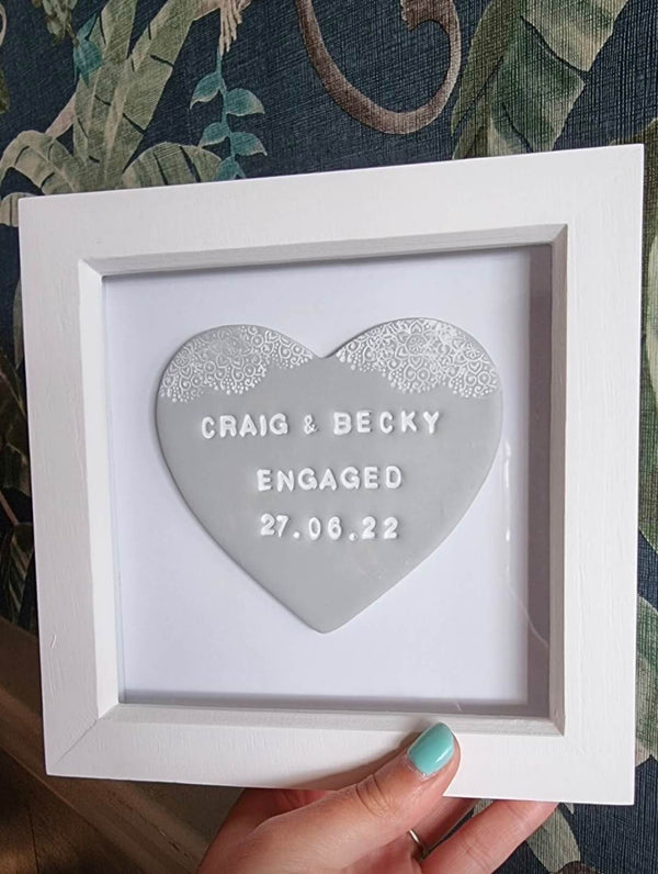 A hand with mint green nail polish holds a white square frame containing a grey polymer clay heart that reads 'CRAIG & BECKY ENGAGED 27.06.22