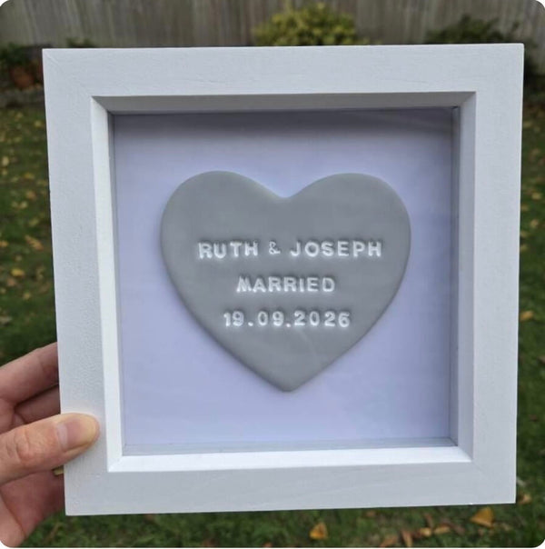 A hand holds a square white shadow box frame containing a grey polymer clay heart with the embossed text 'RUTH & JOSEPH MARRIED 19.09.
