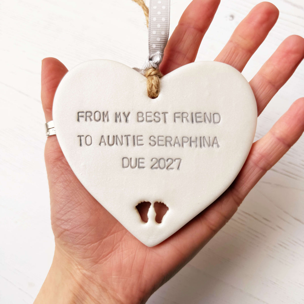 Pearlised white clay hanging heart with baby feet cut out of the bottom and grey personalisation, the heart is personalised with FROM MY BEST FRIEND TO  AUNTIE SERAPHINA  DUE 2027