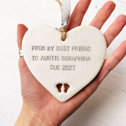 Pearlised white clay hanging heart with baby feet cut out of the bottom and grey personalisation, the heart is personalised with FROM MY BEST FRIEND TO  AUNTIE SERAPHINA  DUE 2027