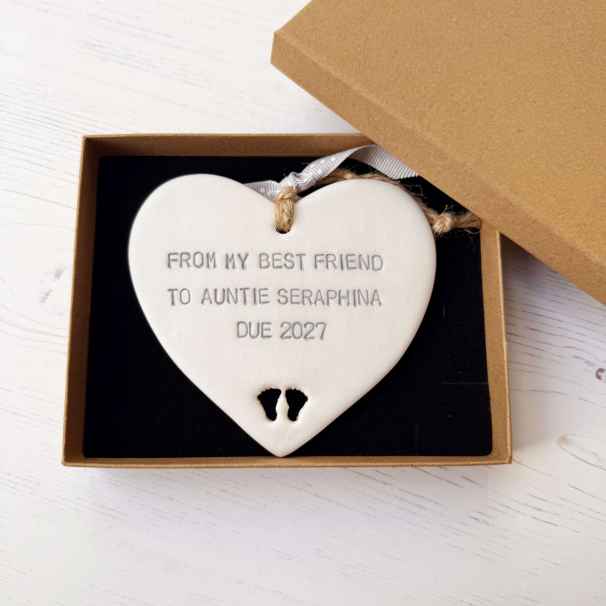 Pearlised white clay hanging heart with baby feet cut out of the bottom and grey personalisation, the heart is personalised with FROM MY BEST FRIEND TO  AUNTIE SERAPHINA  DUE 2027