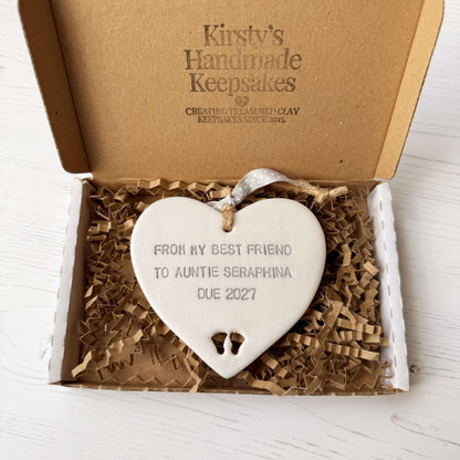Pearlised white clay hanging heart with baby feet cut out of the bottom and grey personalisation, the heart is personalised with FROM MY BEST FRIEND TO  AUNTIE SERAPHINA  DUE 2027