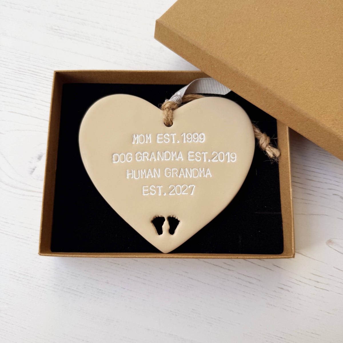 Beige clay hanging heart with baby feet cut out of the bottom and white personalisation, the heart is personalised with MOM EST. 1999 DOG  GRANDMA EST. 2019 HUMAN GRANDMA EST. 2027