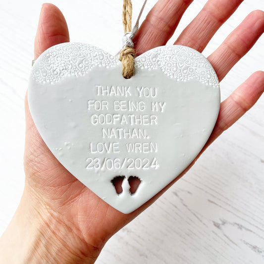 Personalised Godfather gift, grey clay hanging heart with baby feet cut out of the bottom and white lace edge at the top of the heart, the heart is personalised with THANK YOU FOR BEING MY GODFATHER NATHAN. LOVE WREN 23/06/2024